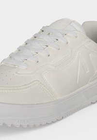 Gros plan d'une basket blanche avec des lacets texturés, des panneaux cousus et un logo embossé sur le côté et la semelle.