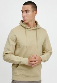 Béžová mikina s kapucí vyrobená z měkkého materiálu, která má přední kapsu, nastavitelné stahovací šňůrky a malé logo na hrudi.