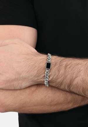 Silver chain bracelet with rectangular black accent. Smooth, shiny links interlock in a thick design. Worn on a wrist with light hair.