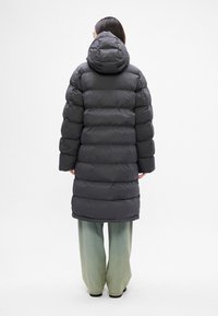 Abrigo de plumas negro con capucha, con costuras en cuadro, silueta hasta la rodilla y mangas largas, combinado con pantalones de pierna ancha en color verde claro.