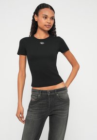 Melnā, ribota īsroku augšdaļa ar apaļu kakla izgriezumu un nelielu logotipu krūšu rajonā, kopā ar pelēkām džinsa biksēm ar priekšējām kabatām.