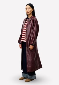 Frau, die einen langen glänzenden burgunderfarbenen Mantel, einen rosa und roten gestreiften Pullover, weite dunkelblaue Jeans und braune Slipper trägt, seitlich stehend.