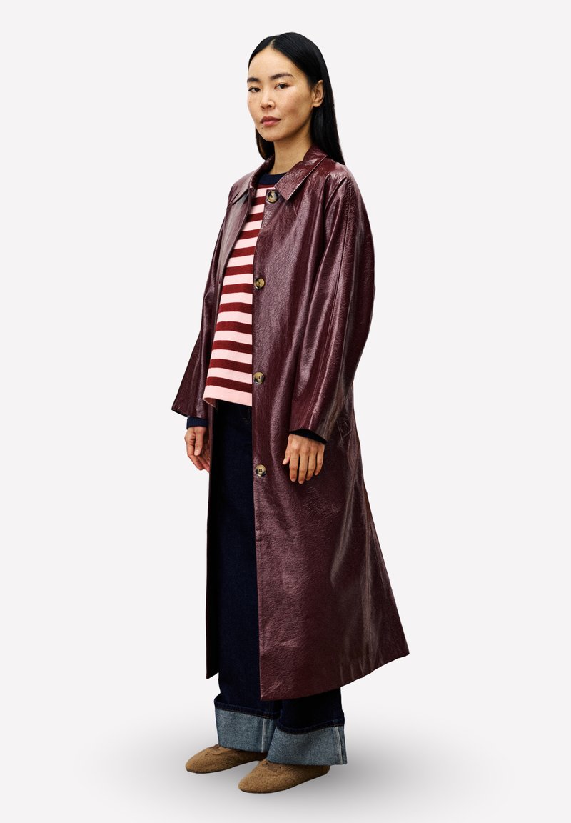 Frau, die einen langen glänzenden burgunderfarbenen Mantel, einen rosa und roten gestreiften Pullover, weite dunkelblaue Jeans und braune Slipper trägt, seitlich stehend.