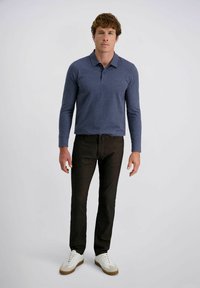Uomo in piedi che indossa un polo a maniche lunghe blu, jeans scuri e sneakers bianche su uno sfondo chiaro.