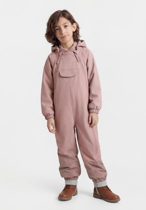 Vaikas su vidutinio ilgio rudos spalvos plaukais, vilkintis dulkiškai rožinį kombinezoną su gaubtu ir rudos spalvos batukus su raišteliais, stovintis prieš baltą foną.