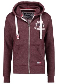 Sudadera con cremallera color marrón burdeos hecha de una mezcla de algodón. Cuenta con capucha, dos bolsillos frontales, cordones blancos y texto decorativo en el pecho.