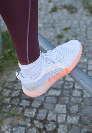 Person iført hvide og koralfarvede Under Armour Phantom løbesko med hvide sokker og burgunder leggings, hviler foden på en metalkrans udendørs.