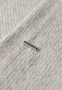 Lys grå tekstureret strikstof med et sølv metalmærke mærket "SUPERDRY" fastgjort, som har to huller til syning.