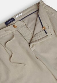 Pantalon beige avec braguette partiellement ouverte, fermeture par bouton et taille à cordon, montrant l’étiquette intérieure "Mr Marvis" et une plus petite étiquette "The Linus."
