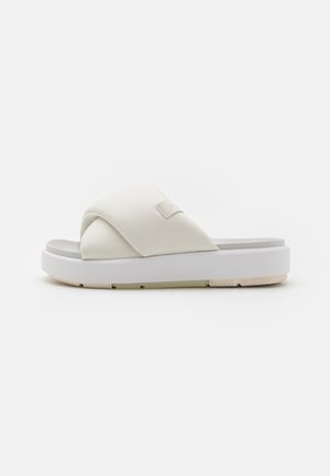 White slide sandals featuring a cross-over strap design, soft texture, and a thick, sturdy sole with detailed tread on the bottom.