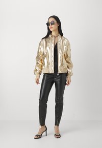 Goldfarbene metallische Bomberjacke mit gerippten Bündchen, schwarze enganliegende Lederhose und schwarze Absatzsandalen. Sonnenbrille und klobige Kettenkette inklusive.