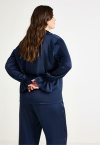 Mörkblå satinblus med samlade klockärmar, med en slät textur och avslappnad passform. Modellen har långt hår och händerna knutna bakom ryggen.