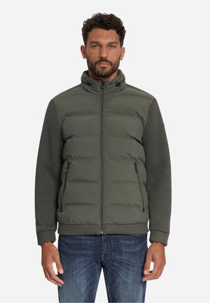 Mann trägt olivgrüne gesteppte Jacke mit gestrickten Ärmeln und Kragen, steht vor weißem Hintergrund mit neutralem Gesichtsausdruck.