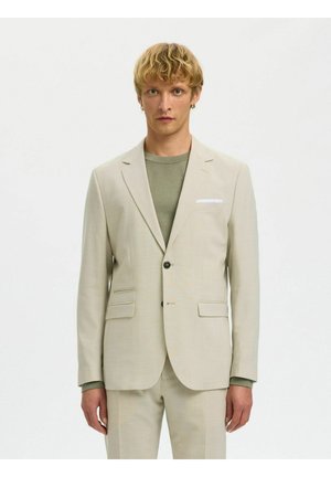 Lyshåret mand iført en beige jakkesætjakke og bukser med en afdæmpet grøn skjorte, stående mod en ensfarvet hvid baggrund.