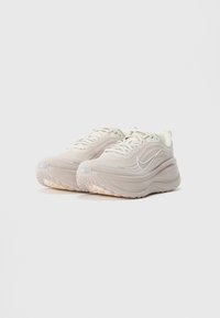 Para beżowych butów biegowych Nike z grubymi, piankowymi podeszwami i sznurowanym fasonem na jednolitym białym tle.
