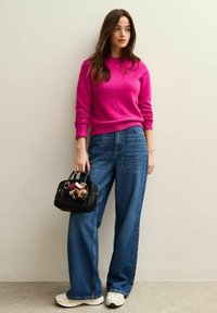 Magenta sweter z ribbami przy mankietach, szerokie niebieskie dżinsy i czarna torebka z pluszowym zawieszką, dopełnione białymi butami sportowymi.