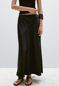 Frau trägt ein schwarzes ärmelloses Oberteil, langen schwarzen Satinsrock, schwarzes Armreif und schwarze flache Sandalen mit offenen Zehen, steht auf weißem Boden.