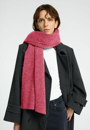 Gebreide roze sjaal met een zachte textuur, gedrapeerd om de nek, gecombineerd met een zwarte jas met een visgraatpatroon en grote knopen.