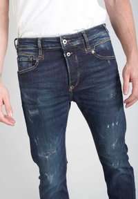 Vaqueros de denim azul oscuro con un corte ajustado, que presentan parches desgastados, un ligero desvanecimiento, herrajes de latón y un diseño clásico de cinco bolsillos.