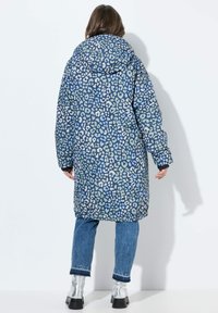 Dlouhá péřová bunda s modro-zeleným leopardím potiskem. Má kapuci a je doplněna modrými džíny a kovově stříbrnými kotníkovými botami.