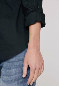 Camicia a maniche lunghe blu scuro con polsino arrotolato e dettaglio con bottone, abbinata a jeans azzurri; mostra una mano appoggiata sulla coscia.