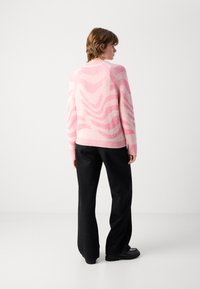 Persoon met de rug naar de camera, gekleed in een roze en witte gebloemde trui, zwarte broek met subtiele stippen en zwarte schoenen, tegen een effen witte achtergrond.