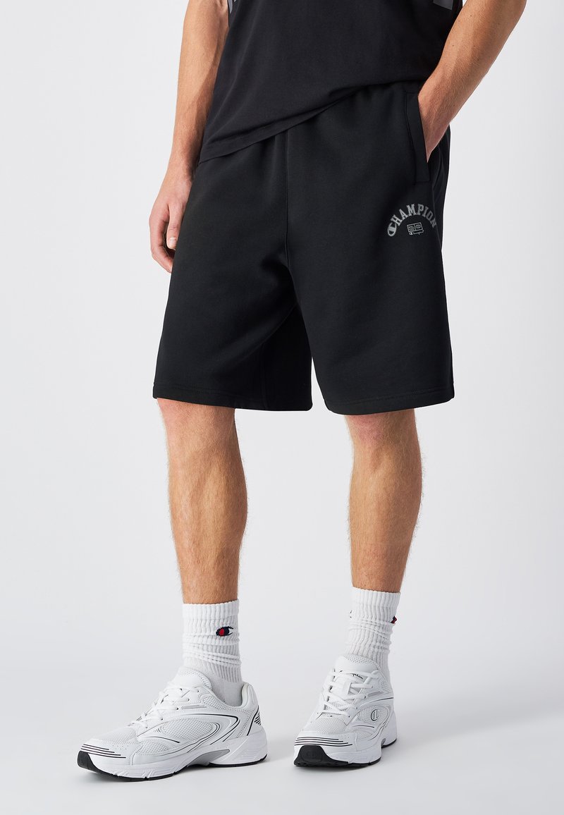Zwarte katoenen shorts met een elastische tailleband en zijzakken. "Champion" logo in het wit gedrukt op het linkerbeen. Gecombineerd met witte sneakers.