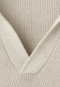 Gros plan d'un tissu beige tricoté montrant un col en V profond avec une texture côtelée et des bords de col pliés.