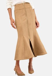 Jupe midi en suédine beige avec fermeture éclair sur le devant, ourlet évasé et coutures verticales. Assortie à des chaussures à talons beiges.