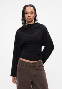 Camiseta negra de manga larga con cuello alto, ajustada en la cintura, combinada con pantalones marrones de corte holgado que cuentan con bolsillos frontales y costura clásica.