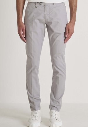 Man draagt lichtgrijze slim-fit broek en witte sneakers, staand tegen een effen achtergrond.