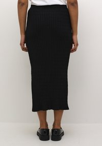 Jupe midi noire en tricot avec une texture côtelée, présentant un design ajusté et un ourlet évasé. Associée à des chaussures noires brillantes.