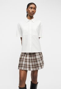 Νεαρή γυναίκα με πλεξούδες, που φοράει λευκό πουκάμισο με κοντά μανίκια και κουμπιά, πλισέ φούστα με καρό σχέδιο και μαύρες μπότες μέχρι το γόνατο, στέκεται μπροστά σε λευκό φόντο.