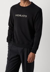 Sweat-shirt noir en tissu doux, avec un col et des poignets côtelés, portant l'inscription "MORATO" en couleur contrastante sur le devant.