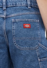 Gros plan sur la poche arrière d'un pantalon en denim bleu avec une étiquette de marque Dickies rouge cousue au centre supérieur de la poche.