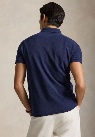 Bărbat văzut din spate, purtând un tricou polo albastru închis cu mâneci scurte și pantaloni bej deschis, stând în fața unui fundal simplu.