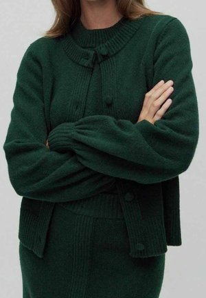 Frau trägt dunkelgrünen Strickcardigan und passenden Rock, Arme verschränkt, zeigt strukturierten Stoff und Details der vorderen Knöpfe.