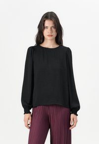 Svart bluse med lange ermer, rund halslinning og rynkede mansjetter, kombinert med plisserte bordeauxbukser. Teksturert overflate på bluse-stoffet.