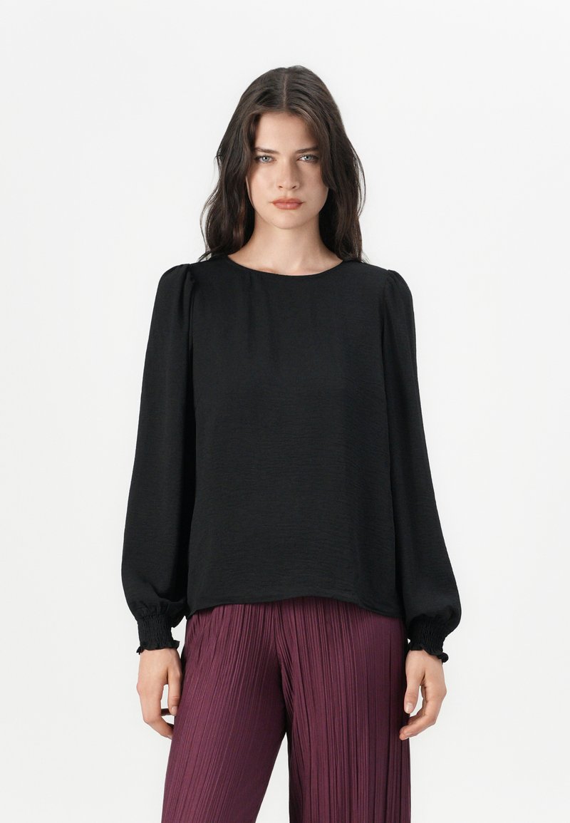 Svart bluse med lange ermer, rund halslinning og rynkede mansjetter, kombinert med plisserte bordeauxbukser. Teksturert overflate på bluse-stoffet.