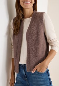 Frau lächelt, trägt eine fusselige braune ärmellose Weste über einem crèmefarbenen Langarmshirt und blauen Jeans, mit einer Hand in der Tasche, vor einer hellen Wand.