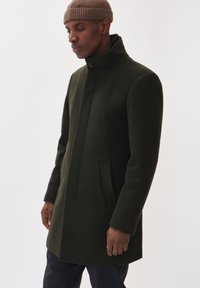 Manteau en laine vert foncé avec un col haut, fermeture à boutons à l'avant et poches latérales. Présente un design épuré et minimaliste avec une texture lisse.