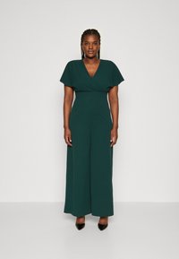 Donkergroene jumpsuit met een wikkel V-hals en korte flutter mouwen, met een getailleerde taille en wijde broekspijpen. Gekozen met zwarte hakken.