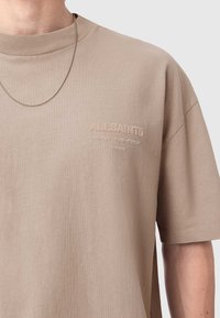 Μπεζ βαμβακερό t-shirt με κοντά μανίκια, με κέντημα που γράφει "ALLSAINTS" και στρογγυλή λαιμόκοψη. Υπάρχει ασημί αλυσίδα κολιέ.