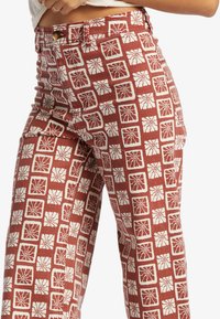 Red patterned pants with white floral designs in square frames, featuring a button closure and a straight leg cut. Cotton fabric with texture.