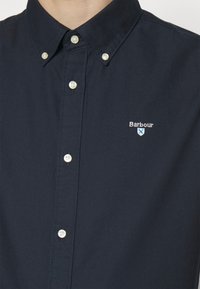 Granatowa koszula z guzikami z klasycznym kołnierzykiem, perłowe białe guziki oraz małe białe haftowane logo "Barbour" na lewej piersi.