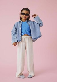 Džínová bunda, modrý texturovaný top, krémové kalhoty s širokými nohavicemi, šedé slip-on boty a sluneční brýle; na růžovém pozadí.