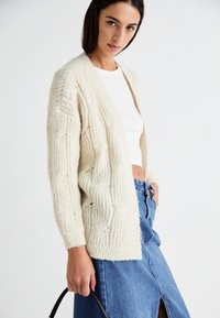Flauschige cremefarbene Strickjacke mit strukturiertem Muster, offenem Vorderteil und langen Ärmeln. Kombiniert mit einem weißen Oberteil und einem blauen Denim-Rock, accessoriert mit einer schwarzen Tasche.