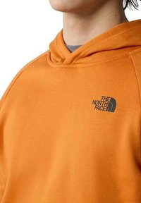 Sudadera con capucha naranja hecha de tela suave, que presenta un logo bordado gris en la parte izquierda del pecho y una capucha con cordón.