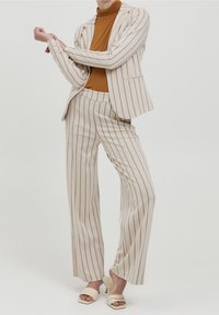 Beżowy garnitur w paski z długimi, szerokimi spodniami i dopasowaną marynarką, noszony na brązowym golfie. Modelka ma na sobie kremowe sandały na obcasie z odkrytymi palcami.