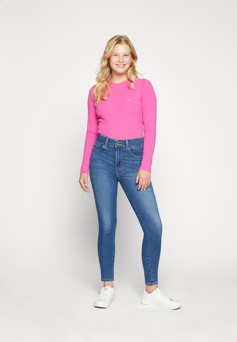 Maglione rosa a costine aderente con maniche lunghe, abbinato a jeans skinny blu a vita alta e sneakers bianche, in piedi su uno sfondo chiaro.